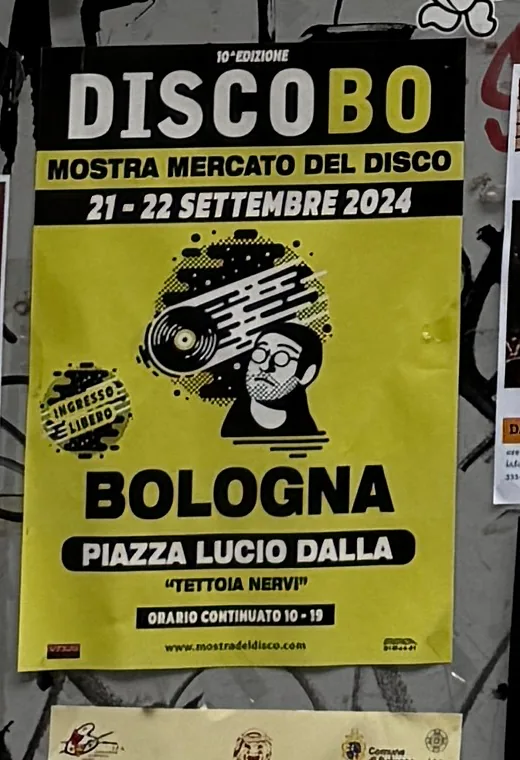 Il volantino giallo della mostra mercato del disco DiscoBo, 21–22 settembre 2024, piazza Lucio Dalla, attaccato alla saracinesca.