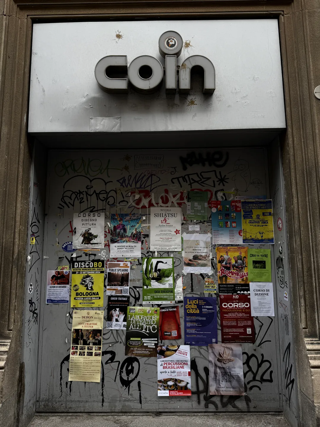 La saracinesca di un ingresso della Coin in via Rizzoli a Bologna: lamiera grigia coperta di graffiti e di più di venti volantini, sopra l'insegna «coin» con le lettere a scatola, il tutto incorniciato dalla pietra del palazzo.
