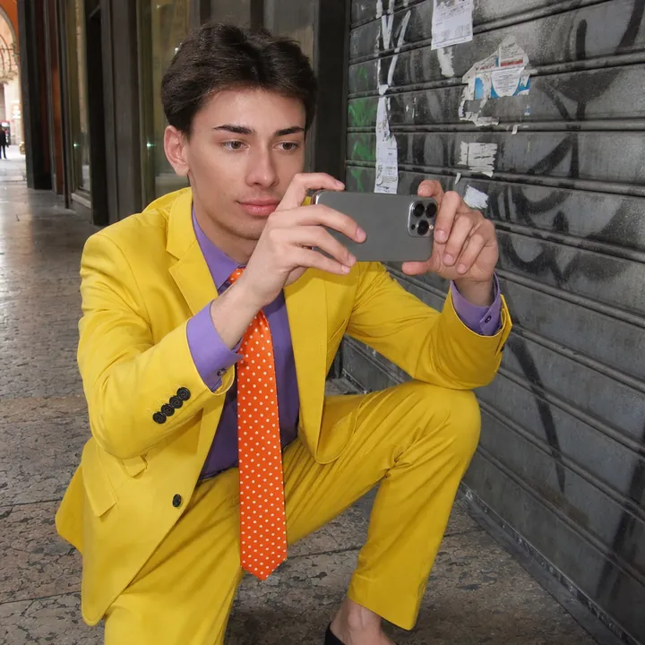 Meme: Pekko in completo giallo si accovaccia come un fotografo di moda e fotografa col telefono nuovo una saracinesca coperta di graffiti.