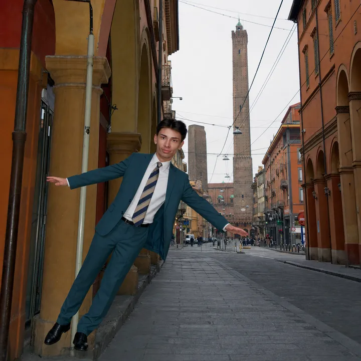 Meme: sotto i portici di Bologna, con le due torri in fondo alla strada, Pekko in completo verde petrolio si inclina di lato come la torre.